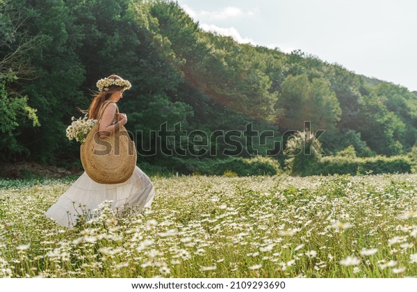 A\
young woman in a white sundress, a wreath of daisies with a large\
wicker bag on her shoulder is walking through a field of daisies,\
against the background of the forest, the sunset\
light
