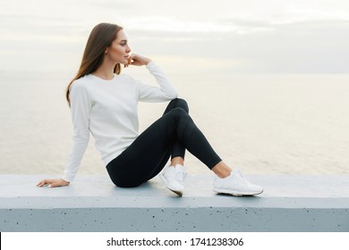 Young woman in white sweater and black leggings is resting after jog on promenade. Lifestyle, beauty, walking outdoors