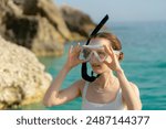 A young woman in a white swimsuit with a transparent mask and a snorkel on her face is going to swim against the background of the sea with rocks. Concept of summer vacation, diving and snorkeling