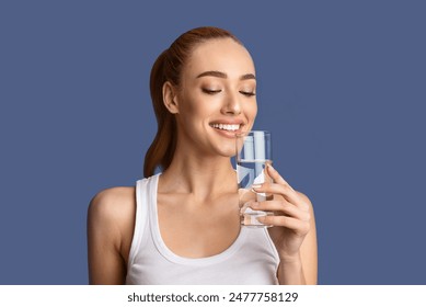 A young woman in a white tank top smiles as she drinks water from a clear glass. She is standing against a solid blue background. - Powered by Shutterstock