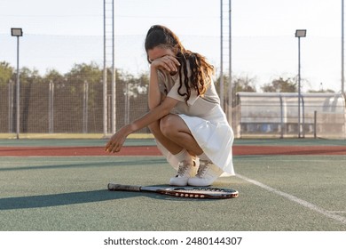 A young woman in a white tennis dress sits on the court, crying after a point during a match - Powered by Shutterstock