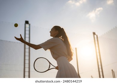 young woman in a white tennis outfit stands on a tennis court, preparing to serve the ball. The sun shines brightly behind her, creating a beautiful silhouette - Powered by Shutterstock