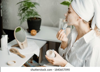 Young woman in white towel chilling in bedroom and making clay facial mask near mirror. Girl doing beauty treatment and relaxing at home. Morning skin care beauty routine, self care