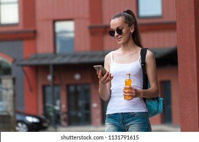 A young woman in a white T-shirt and black sunglasses goes home after training and is holding a mobile phone and an orange cocktail in her hand. Copy space