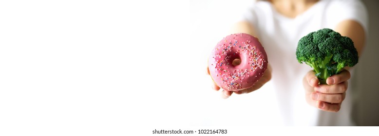 Young Woman In White T-shirt Choosing Between Broccoli Or Junk Food, Donut. Healthy Clean Detox Eating Concept. Vegetarian, Vegan, Raw Concept. Copy Space. Banner.