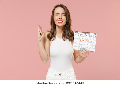 Young woman in white t-shirt hold female periods calendar checking menstruation day wait special moment keep finger cross make wish isolated on pink background Medical healthcare gynecological concept