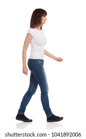 Young Woman In White T-shirt, Jeans And Sneakers Is Walking And Looking Away. Side View. Full Length Studio Shot Isolated On White.