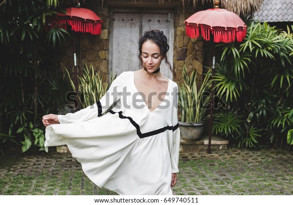 Young woman in white tunic in\
Ubud village with traditional balinese architecture. Style of Bali\
house. Fashion style, curly hair, light dress. Villa in\
Changgu