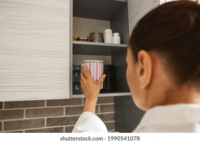 Young Woman In A White Waffle Dressing Gown Opens A Cupboard In The Kitchen And Takes Out A Jar Of Coffee. Rear View. Morning Routine. Breakfast Time