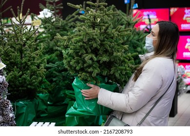 Young Woman In White Winter Jacket And Protective Mask In Shopping Center Chooses A Real Christmas Tree To Decorate House For Holidays. Christmas . Personal Protective Equipment.