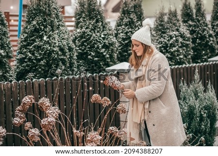 Young woman in winter clothes and knitted white hat with scarf in the winter garden chooses snow - covered hydrangeas for New Year 's decor . Snow is falling. Garden in winter. Frozen plants.