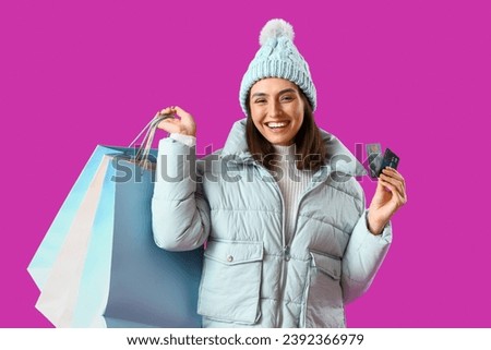 Young woman in winter clothes with shopping bags and credit cards on purple background