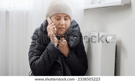 Young woman in winter coat and hat sitting by the broken heater in freezing home and calling repair service.