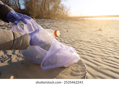 A young woman in a winter jacket holds a garbage bag on a river or sea beach at sunset. Highlighting environmental cleanup efforts, the warm sunset light contrasts with the pollution challenge - Powered by Shutterstock