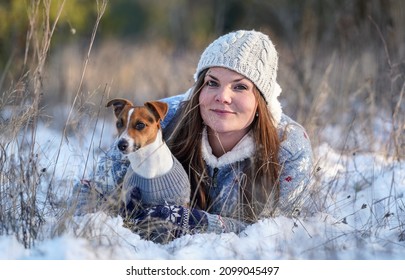 Young woman in winter jacket lying down at snow covered ground, holding her Jack Russell terrier dog, blurred trees background - Powered by Shutterstock