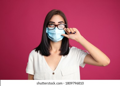 Young woman wiping foggy glasses caused by wearing disposable mask on pink background. Protective measure during coronavirus pandemic