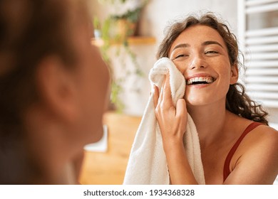 Young woman wiping her face with towel after waking up in the morning. Beautiful happy smiling girl holding towel near facial skin after washing face. Happy woman cleaning and drying skin with napkin.