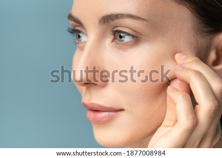 Young woman without makeup touching cheeks after glycolic acid peel, has signs of aging skin on her face, close up, isolated on studio blue background with copy space. 