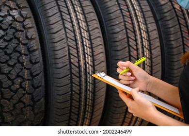 A Young Woman Worker With A Clipboard Checking Stocks Of New Tires Ready To Be Replaced At A Service Center Or Tire Shop. The Background Is A Warehouse. Stock New Tires For The Car Industry.