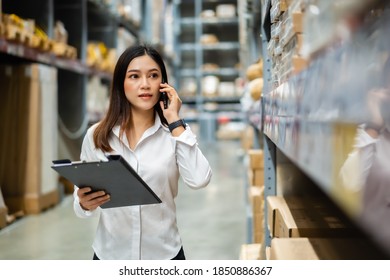 Young Woman Worker Talking On A Mobile Phone And Holding Clipboard To Checking Inventory In The Warehouse Store