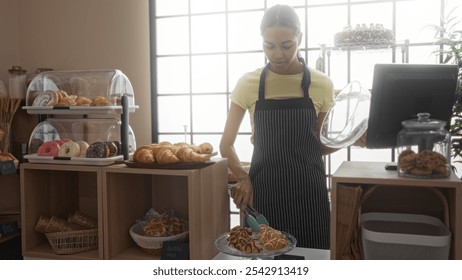 Young woman working in a bakery, arranging pastries and cakes, in an indoor shop with natural light from a large window - Powered by Shutterstock