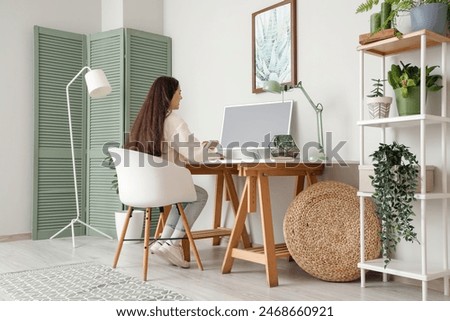Young woman working with computer and hanging painting on light wall in office