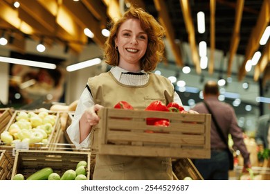 Young woman working in grocery store carrying wooden crate full of fresh red bell peppers, smiling at customers in produce section - Powered by Shutterstock