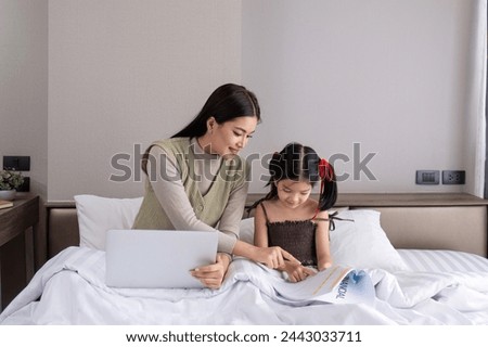 A young woman is working from home with her adorable daughter, watching work together and happily teasing each other in the bedroom.