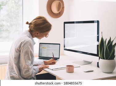 Young woman working at home office. Girl wearing in glasses works with new startup project. Female thinking and writing in planner or diary at workplace of minimalist style with computer.