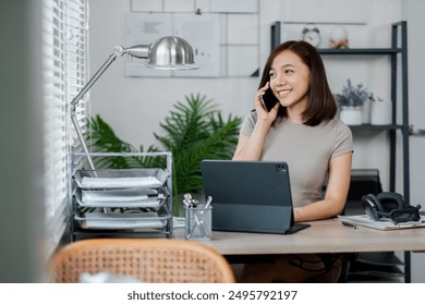 Young woman working from home office, smiling while talking on phone and using tablet, modern and organized workspace. - Powered by Shutterstock