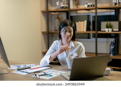 A young woman is working late at home and eating instant noodles for dinner - Powered by Shutterstock