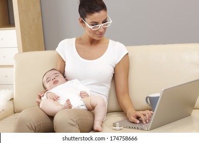 Young woman working on laptop computer while holding sleeping baby in arms.