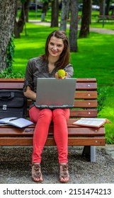Young woman working on a laptop outside in a park oferring a green apple and looking to the camera. The main focus is on the apple.
