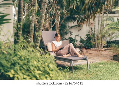 Young woman working on laptop in the house garden.Young woman lies on a sun lounger in the garden and works on her laptop. Digital nomad concept.