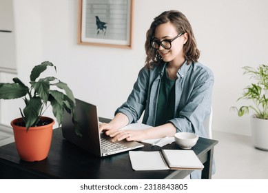 Young woman working on laptop in professional office setting. Student girl multitasking at home office. Work-from-home, freelance, business lifestyle concept. Technology, productivity, and remote work