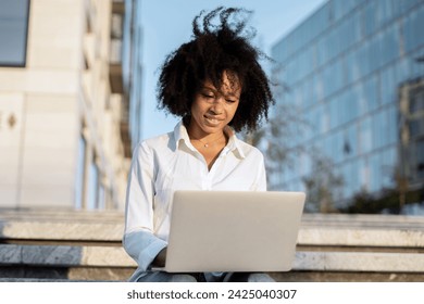Young woman working on laptop outdoors, urban setting, displaying focus and casual professionalism.