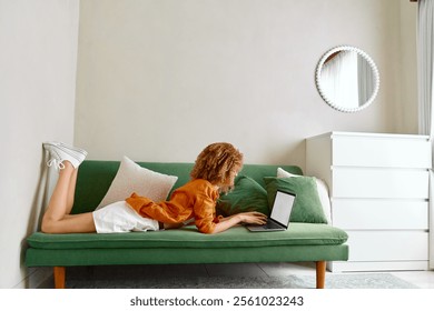 Young woman working on a laptop at home, relaxed on a green sofa, casual outfit, showcasing productivity in a modern interior - Powered by Shutterstock