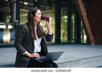 Young woman working online on laptop computer sitting outside the office building wearing wireless headphones enjoying a takeaway coffee. Freelancer working