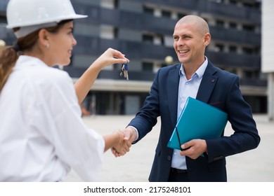 Young Woman Working As A Process Engineer At A Construction Site, With A Polite Smile, Hands Over The Keys To An Flat In A ..new Building To A Man Customer