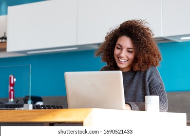 Young woman working remotely from her home kitchen, sitting at the counter with laptop and smiling, looking at computer screen. Low angle portrait with copy space