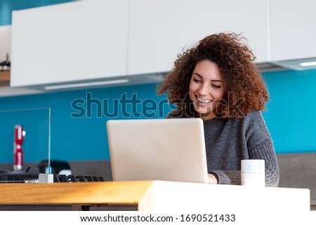 Young woman working remotely from her home kitchen, sitting at the counter with laptop and smiling, looking at computer screen. Low angle portrait with copy space
