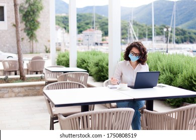 Young Woman Working Safety In Empty Cafe Outdoors. Social Distancing During Quarantine. Freelancer Wearing Protective Mask, Using Laptop. Lifestyle Moment. Restaurant Patio With Safety Limited Seating