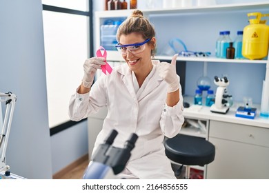 Young Woman Working At Scientist Laboratory Holding Pink Ribbon Smiling Happy And Positive, Thumb Up Doing Excellent And Approval Sign 