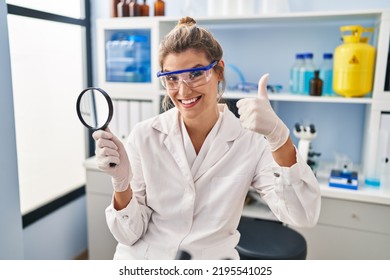 Young Woman Working At Scientist Laboratory Holding Magnifying Glass Smiling Happy And Positive, Thumb Up Doing Excellent And Approval Sign 