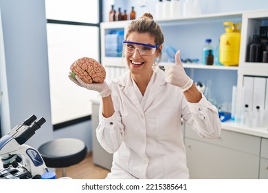 Young Woman Working At Scientist Laboratory Holding Brain Smiling Happy And Positive, Thumb Up Doing Excellent And Approval Sign 