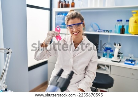 Young woman working at scientist laboratory holding pink ribbon looking positive and happy standing and smiling with a confident smile showing teeth 