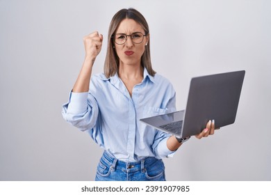 Young woman working using computer laptop angry and mad raising fist frustrated and furious while shouting with anger. rage and aggressive concept. 
