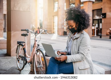 Young woman works at laptop sitting on a bench to send urgent work on the way to home with her bicycle after a day of work, wearing protective face mask during the global Coronavirus Covid-19 pandemic