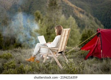 Young Woman Works On Laptop While Sitting Relaxed On Chair By The Campfire, Traveling With Tent In The Mountains. Concept Of Remote Work And Escape To Nature