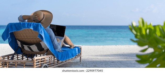 A young woman works on a laptop on the beach in the Maldives. The concept of remote work and nomadism. Back view. Banner - Powered by Shutterstock